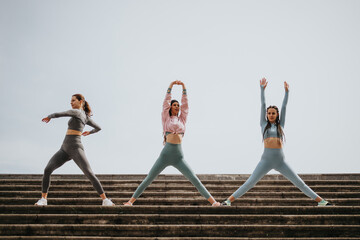 Three fit women in athletic wear practicing yoga, stretching outdoors against a clear sky...