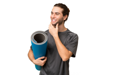 Young sport man going to yoga classes while holding a mat over isolated background looking to the side and smiling