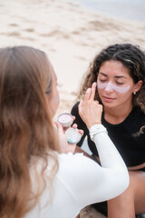 A young blonde woman applies sun protective surfer zinc to the face of her Latino woman friend with curly hair who is sitting opposite her. Skin protection for surfers. Waterproof protective zinc.