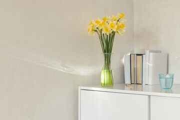 Bouquet of yellow spring flowers on the white dresser against empty wall. Books and flowers indoors interior.Empty space