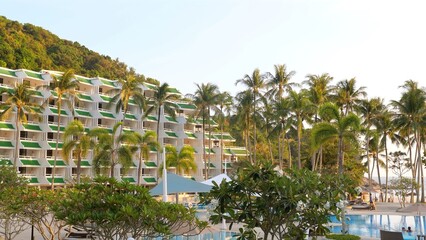 Tropical resort hotel with large swimming pool at forefront surrounded by lush palm trees and hillside full of holiday apartments. Travel and luxury accommodation.