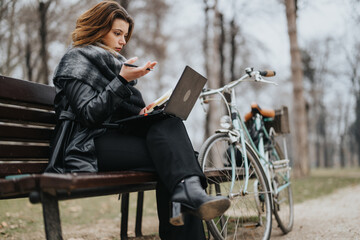 Fototapeta na wymiar Stylish young woman confidently working on a laptop while sitting on a bench in a park, with a vintage bicycle beside her.