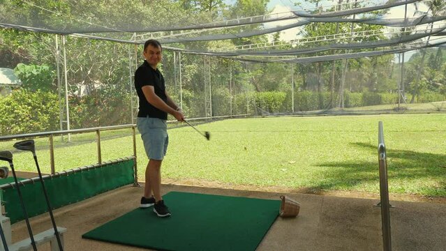 Focused man practicing golf swing on driving range with greenery in background. Outdoor recreational activities and sports training.