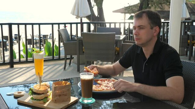 Man enjoying meal with pizza and sandwich at beachside restaurant, with tropical ocean view in background. Relaxation and vacation dining experience.