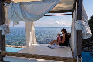 Woman holding a baby in lounger canopy beach bed in luxury hotel with scenic view of idyllic...