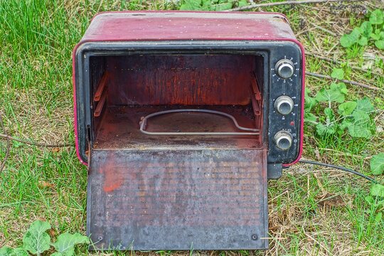 one old rusty iron cherry broken discarded electric powerful open with a timer microwave oven stands on the green grass on the street during the day