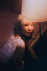 Portrait of a beautiful Asian woman with blonde hair and white wings, illuminated by the soft light of a floor lamp in the evening. She looks like an angel.
