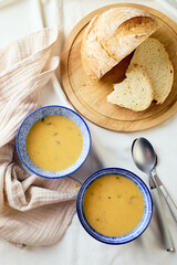 Two bowls of creamy potato soup, spoons, napkin and wooden cutting board with a loaf of bread and cut pieces.