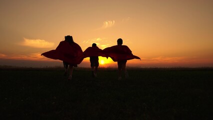 Group of children dressed as superheroes runs across meadow against of setting sun. Silhouette of...