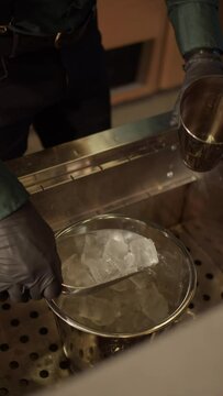 Bartender pouring ice from scoop into cocktail shaker, ice cubes falling. Bartender puts ice cubes in a shaker with a bucket close-up. Vertical video.