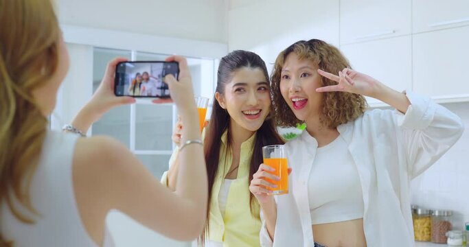 Two smiling women posing for a selfie with refreshing drinks as a friend takes their photo in a sunny kitchen.