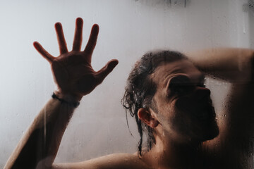 A contemplative man behind a foggy shower door presses his hand to the glass, partially obscuring his face.