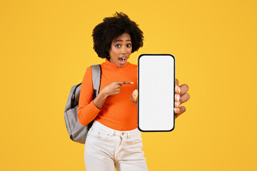 Excited young woman with curly hair, pointing at a blank smartphone screen, dressed in an orange...