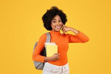 Cheerful young woman with afro hair holding notebooks, making a call me gesture, clad in an orange...