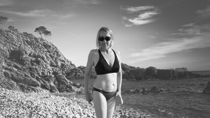 woman with black sunglasses and hat walking on the sandy beach. black and white image - obrazy, fototapety, plakaty