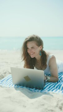 happy stylish middle aged woman on the beach with straw bag, laptop and striped towel.