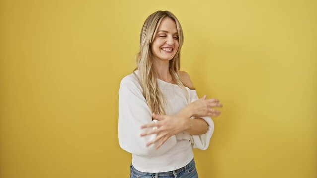 Joyful young blonde woman standing, arms raised in victory, celebrating successful win, eyes closed in happiness over isolated yellow background