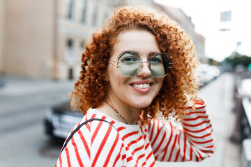 Outdoor portrait of stylish ginger head girl in odd-shaped sunglasses walking down big city streets, visiting popular touristic places, turning to camera, petting her curly hair with shy smile - 779143592