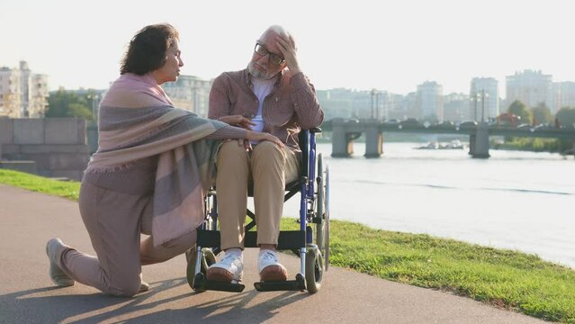 Old man in wheelchair walking with caregiver senior woman in park. Elderly family couple woman supporting taking care of paralyzed man in chair for people with disability outdoor. Rehabilitation
