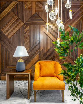 Modern orange armchair, lamp shade on small wooden table, planter with green bushes, and contemporary tall glass chandelier, in a hall with decorated wood cladding wall, and white marble floor