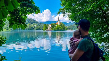 Man holding baby with scenic view of St Mary Church build on small island on alpine lake Bled, Upper Carniola, Slovenia. Serene landscape in Julian Alps in winter. Hills covered with lush green forest