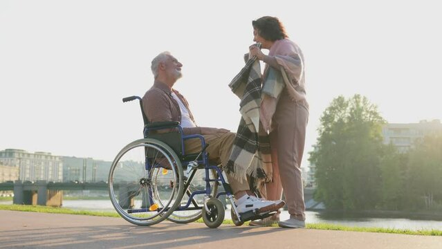 Old man in wheelchair walking with caregiver senior woman in park. Elderly family couple woman supporting taking care of paralyzed man in chair for people with disability outdoor. Rehabilitation