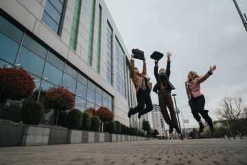 Three exuberant young business professionals leap into the air, celebrating success outside a modern office building.