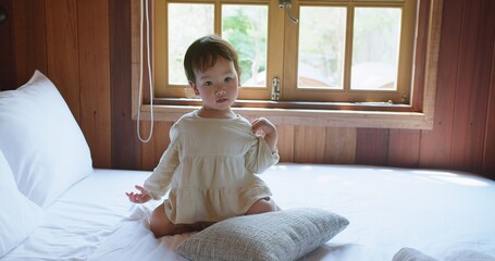 A young Asian child girl in a white robe sits on a large bed relaxing resting in bedroom, gazing...