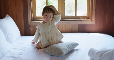 A young Asian child girl in a white robe sits on a large bed relaxing resting in bedroom, gazing...