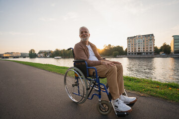 Happy senior man in wheelchair smiling enjoying freedom walking on road in city park outdoor. Old...