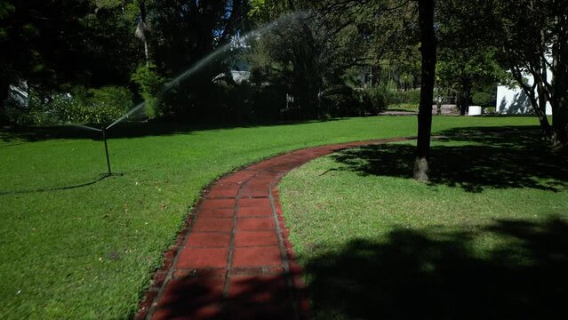 A sprinkler system is watering a lush, green lawn adjacent to a red brick pathway