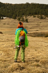 A woman with green hair is walking in a field with a backpack on her back. The backpack is green and has a black strap. The woman is wearing a blue shirt. The field is dry and brown