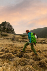 Naklejka premium A woman is walking on a hill with a green backpack. The sky is orange and the grass is brown