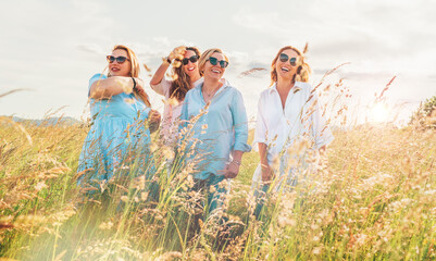 Portrait of four cheerful smiling and laughing women during outdoor walking by a high green grass hill. They looking at the camera. Woman's friendship, relations, and happiness concept image.