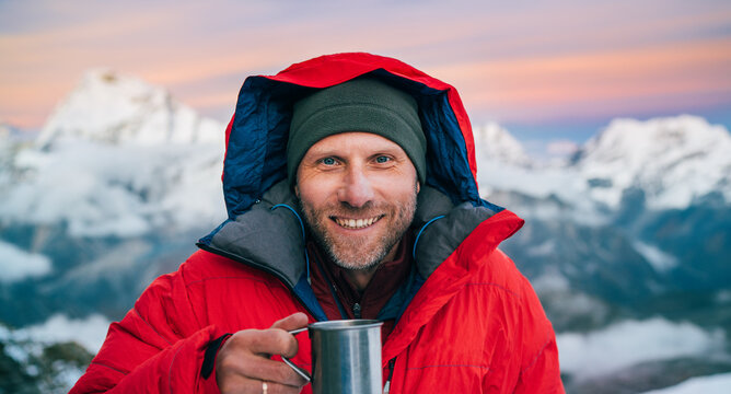 Portrait of smiling at camera high altitude mountaineer dressed red warm dawn jacket holding metal mug of hot tea in with mountains panorama background. Mera Peak High Camp 5700m, Himalayas, Nepal.