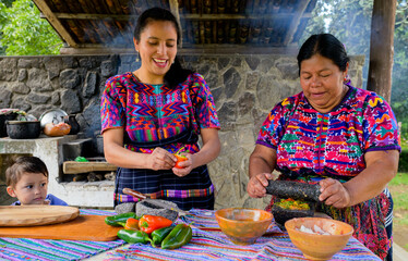 A boy watches as his mother and grandmother from the Mayan ethnic group grind the vegetables using...