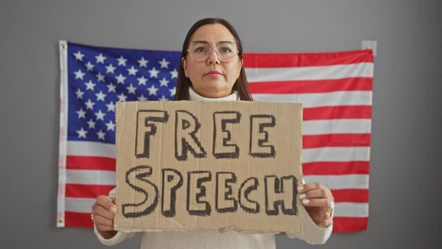 A middle-aged woman stands indoors holding a 'free speech' sign before an american flag.