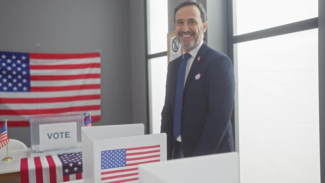 A mature man with a beard smiles in a voting booth at an american polling station, adorned with flags and 'vote' signage.