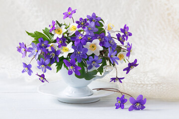 Bouquet of spring flowers hepatica and anemones in a cup on a white wooden table, beautiful card - 779219139