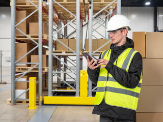 Man warehouse contractor. Guy with phone in storage area. Warehouse racks with boxes behind storekeeper. Contractor courier storage. Worker in helmet holds phone. Man logistics warehouse contractor