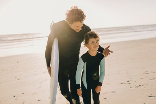 Father and son in wetsuits walking on the beach with a surfboard