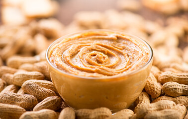Peanut butter in a glass bow over raw peanuts background. Creamy smooth peanut butter in glass bowl backdrop. Texture. Organic food. American cuisine. 