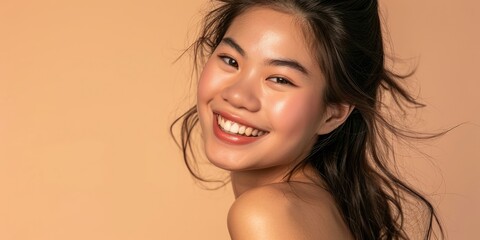 A stunning portrait of a radiant Asian woman with a beaming smile and glowing skin against a soft peach background, epitomizing natural skincare beauty..