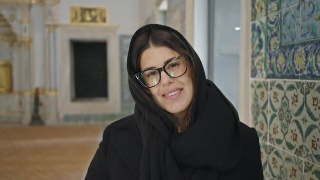Smiling woman with glasses and headscarf poses in historical topkapi palace, istanbul, exuding charm and cultural immersion.