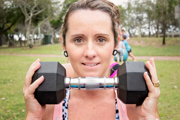 Woman in fitness gear holding dumbbell, looking straight at camera, closeup, outdoors in park