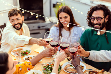 Group of young friends drinking red wine enjoying summer barbecue at rooftop terrace. Diverse people having fun at dinner party