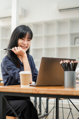 Cheerful young woman with pen in hand working on her laptop in a bright cafe, with a coffee cup on the table.