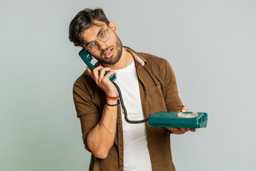 Tired bored Indian man talking on wired vintage telephone of 80s, fooling, making silly faces, exhausted of tedious story, not interested in communication talk. Arabian guy isolated on gray background