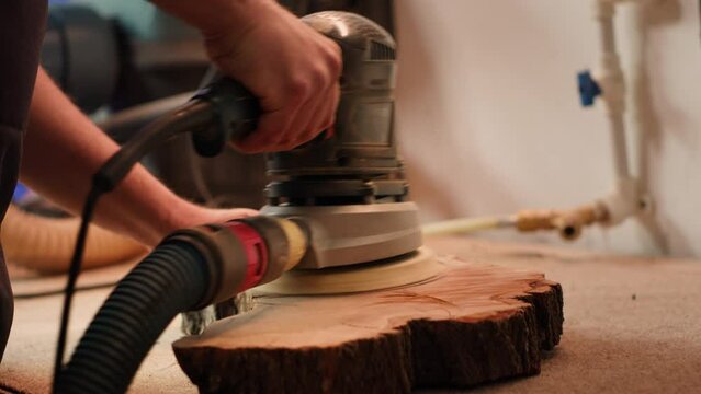 Craftsperson works with orbital sander on lumber block to achieve smooth finish, close up shot. Woodworking specialist in furniture assembly shop using angle grinder to fix wooden surface, camera A