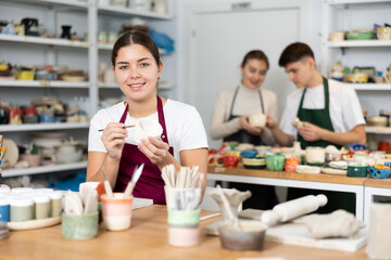 Smiling young woman in apron painting on handmade clay cup with brush in pottery workshop. Persons...